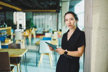 Elinde tabletle siyah takım elbiseli tatlı bir kadın modern bir ofiste duruyor..