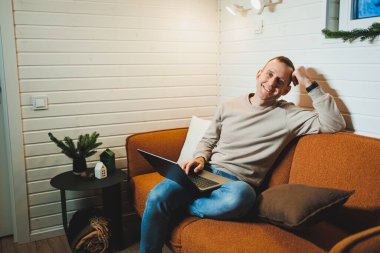 Ev kıyafetleri içinde yakışıklı bir adam kanepeye oturur ve dizüstü bilgisayarda çalışır. Uzak serbest çalışma.