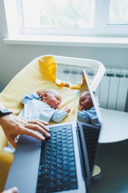 Genç bir baba dizüstü bilgisayarda çalışıyor ve bir bebeğin yanında oturuyor. Evde küçük bir çocukla çalışmak.