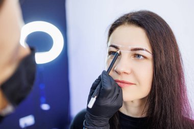 Usta, kalıcı kaş makyajından önce bir kadının yüzünü işaretler. Kaş dövmesi. Bir kadının yüzü için estetik ameliyat