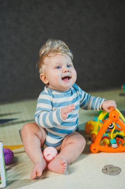 7 aylık bir çocuğun yerde oturup oyuncaklarla oynadığı fotoğraf. Çocuk gülümsüyor ve oyuncaklarla oynuyor..