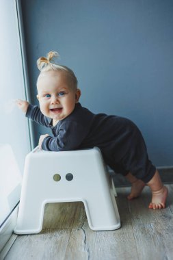 Evin içinde 7-9 aylık bir çocuğun fotoğrafı. Meraklı, gülümseyen bir çocuk etrafındaki dünyayı keşfediyor. Sandalyenin yanında duran sevimli bebek. Çocuk ayağa kalkıp yürümeyi öğreniyor..