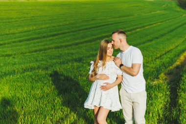 Yeşil bir bahar tarlasında sarılan hamile bir çift. Beyaz çiçek açan ağaçların parkında beklenen ebeveynler. Bebek bekleyen romantik bir çift. Yeşil bir tarlada yürüyüş