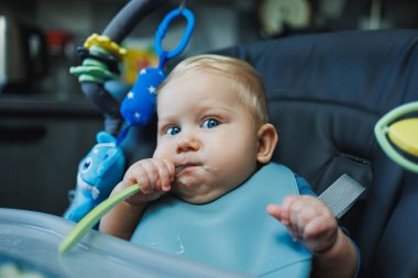 Bebek yüksek sandalyede oturan örülü süveter giyiyor ve acıkıyor, kaşık tutuyor ve püre veya lapa yiyor, sarı şişede elma ve suyun tadını çıkarıyor, beyaz arka planda izole bir şekilde poz veriyor..