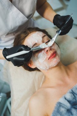 Tıp ve kozmetoloji. Siyah eldivenli bir güzellik uzmanının elleri bir kadının yüzüne maske takar. Kişisel cilt bakımı. Yüze nemlendirici soyulan maske.