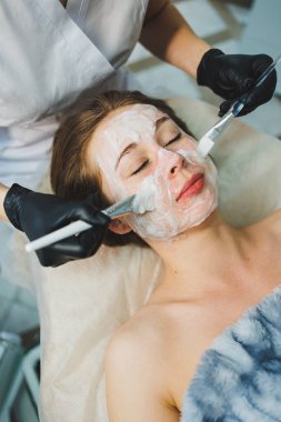 Tıp ve kozmetoloji. Siyah eldivenli bir güzellik uzmanının elleri bir kadının yüzüne maske takar. Kişisel cilt bakımı. Yüze nemlendirici soyulan maske.