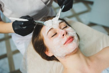 Tıp ve kozmetoloji. Siyah eldivenli bir güzellik uzmanının elleri bir kadının yüzüne maske takar. Kişisel cilt bakımı. Yüze nemlendirici soyulan maske.