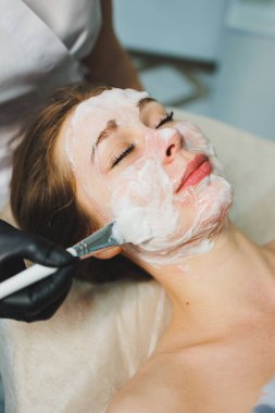 Tıp ve kozmetoloji. Siyah eldivenli bir güzellik uzmanının elleri bir kadının yüzüne maske takar. Kişisel cilt bakımı. Yüze nemlendirici soyulan maske.