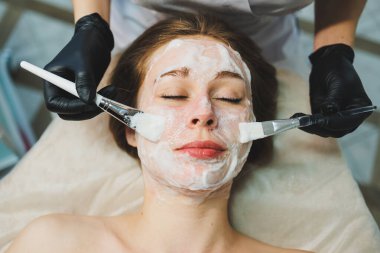 Tıp ve kozmetoloji. Siyah eldivenli bir güzellik uzmanının elleri bir kadının yüzüne maske takar. Kişisel cilt bakımı. Yüze nemlendirici soyulan maske.