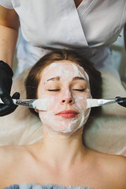 Tıp ve kozmetoloji. Siyah eldivenli bir güzellik uzmanının elleri bir kadının yüzüne maske takar. Kişisel cilt bakımı. Yüze nemlendirici soyulan maske.