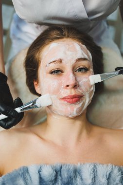 Tıp ve kozmetoloji. Siyah eldivenli bir güzellik uzmanının elleri bir kadının yüzüne maske takar. Kişisel cilt bakımı. Yüze nemlendirici soyulan maske.