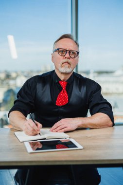 Siyah gömlekli gri saçlı bir adam ofiste bir masada oturuyor ve bir tabletle çalışıyor. Modern çalışma alanı