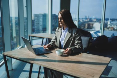 Ofiste çalışırken elinde bir fincan kahve ve dizüstü bilgisayarla masada oturan genç bir yönetici kadın..