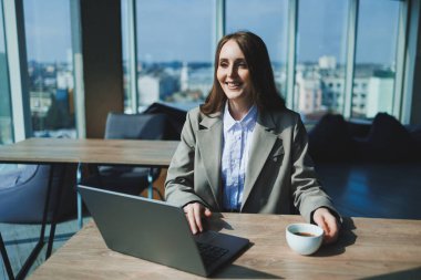 Ofiste çalışırken elinde bir fincan kahve ve dizüstü bilgisayarla masada oturan genç bir yönetici kadın..