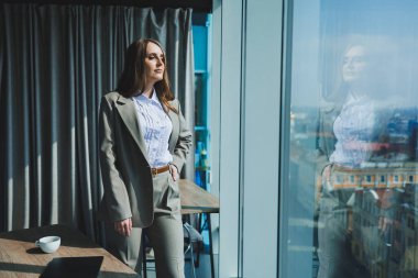 Klasik takım elbiseli ve beyaz gömlekli bir iş kadını modern bir ofiste pencerenin önünde duruyor. Ofisteki masadaki modern iş kadını.