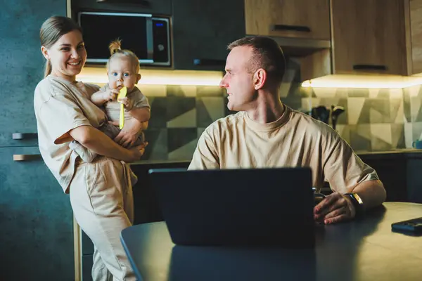 stock image A young couple with a child in the kitchen. A man works at home on a laptop, and his wife with a child in her arms is standing next to him. Family relationships