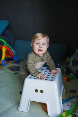 Mavi gözlü ve sarı saçlı küçük bir erkek bebek bir sandalyenin yanında duruyor. Sevimli duygusal bir bebek ayakları üzerinde durmayı öğreniyor.