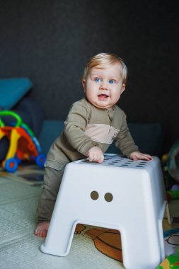 Mavi gözlü ve sarı saçlı küçük bir erkek bebek bir sandalyenin yanında duruyor. Sevimli duygusal bir bebek ayakları üzerinde durmayı öğreniyor.