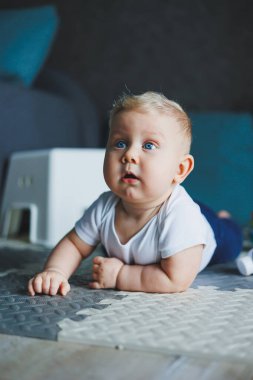Beyaz tulumlu küçük bir çocuk temiz ve sıcak zeminde sürünmeyi öğreniyor. Sevimli mavi gözlü bir bebeğin ev fotoğrafı..