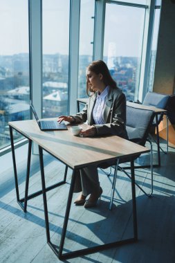 Ofiste çalışırken elinde bir fincan kahve ve dizüstü bilgisayarla masada oturan genç bir yönetici kadın..