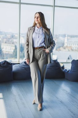 Klasik gri takım elbiseli ve beyaz gömlekli bir iş kadını modern bir ofiste pencerenin önünde duruyor. Ofisteki masadaki modern iş kadını.