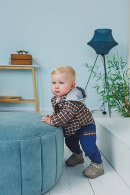 9 aylık şık gömlekli, pantalonlu ve spor ayakkabılı bir erkek bebek bir desteğin yanında duruyor..
