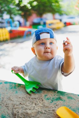 Mavi şapkalı ve tişörtlü küçük bir çocuk kumda plastik bir tırmıkla oynuyor. Oyun bahçesinde bir bebek oynuyor..