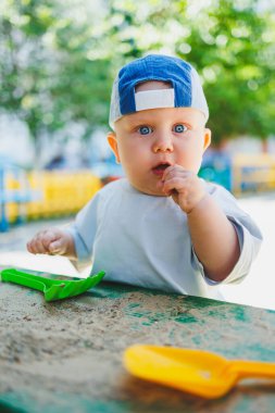 Mavi şapkalı ve tişörtlü küçük bir çocuk kumda plastik bir tırmıkla oynuyor. Oyun bahçesinde bir bebek oynuyor..