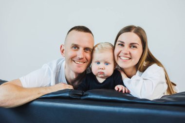 Küçük çocuklu genç bir çift, beyaz bir arka planda bir kanepede otururken fotoğraf çekiliyor. İlgili ebeveynler bebeklerini kollarında tutarken onlara bakarlar..