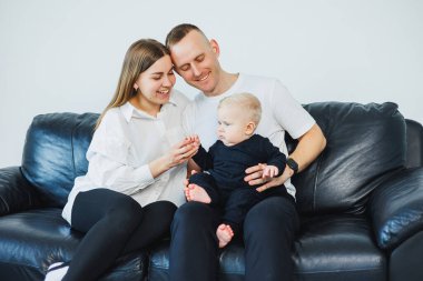 Küçük çocuklu genç bir çift, beyaz bir arka planda bir kanepede otururken fotoğraf çekiliyor. İlgili ebeveynler bebeklerini kollarında tutarken onlara bakarlar..