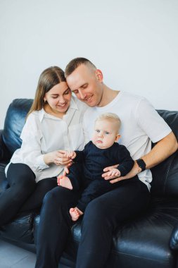 Küçük çocuklu genç bir çift, beyaz bir arka planda bir kanepede otururken fotoğraf çekiliyor. İlgili ebeveynler bebeklerini kollarında tutarken onlara bakarlar..
