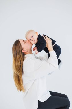 Genç bir anne beyaz arka planda oturur, bebeğini kollarına alır ve onu öper. Anne ve küçük oğlu..