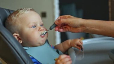 Küçük bir çocuk yüksek bir sandalyede oturuyor, yulaf lapası yiyor ve annesi onu kaşıkla beslemeye çalışıyor. Bebek maması. Bir çocuk için ilk yemek.