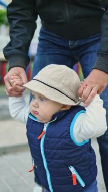 Bir bebek sokakta annesinin elini tutarak yürümeyi öğrenir. Bebeğin ilk adımları. Bebeğin gelişimi. Bebekler için kıyafetler. Dikey video