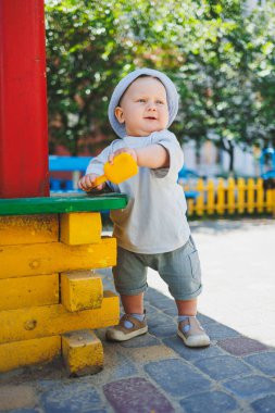 11 aylık, tişörtlü ve kafasında panama olan küçük bir erkek bebek, bir yaz günü kumla oynuyor. Sokakta bir çocuk oynuyor..