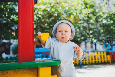 11 aylık tişörtlü ve kafasında panama olan bir erkek bebek bir yaz günü kumla oynar. Sokakta küçük bir çocuk oynuyor.