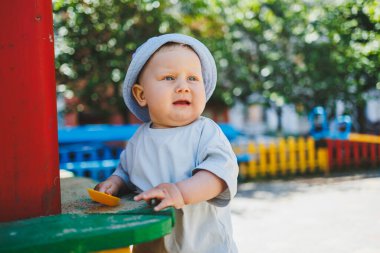 11 aylık tişörtlü ve kafasında panama olan bir erkek bebek bir yaz günü kumla oynar. Sokakta küçük bir çocuk oynuyor.