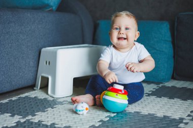 Beyaz elbiseli bir erkek bebek evde bir halıya oturup plastik bir piramitle oynuyor. Bebekler için eğitici oyunlar