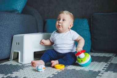 Beyaz elbiseli bir erkek bebek evde bir halıya oturup plastik bir piramitle oynuyor. Bebekler için eğitici oyunlar