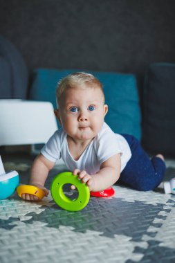 Beyaz elbiseli bir erkek bebek evde bir halıya oturup plastik bir piramitle oynuyor. Bebekler için eğitici oyunlar