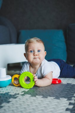 Beyaz elbiseli bir erkek bebek evde bir halıya oturup plastik bir piramitle oynuyor. Bebekler için eğitici oyunlar