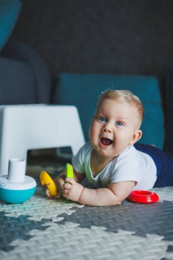 Beyaz elbiseli bir erkek bebek evde bir halıya oturup plastik bir piramitle oynuyor. Bebekler için eğitici oyunlar