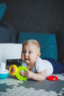Beyaz elbiseli bir erkek bebek evde bir halıya oturup plastik bir piramitle oynuyor. Bebekler için eğitici oyunlar