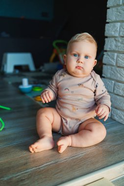 Açık renk saçlı, mavi gözlü bir çocuk. Bebek, 11 ay boyunca mayoyla evde oturuyor ve gülümsüyor. Çocuklar evde oynuyorlar..