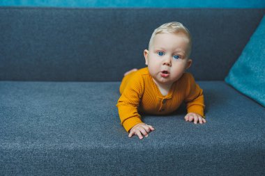 Sarı takım elbiseli sevimli, sarı saçlı küçük bir çocuk kanepede oturuyor. Bebekler için kıyafetler..