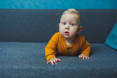 Sarı takım elbiseli sevimli, sarı saçlı küçük bir çocuk kanepede oturuyor. Bebekler için kıyafetler..