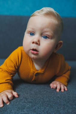 Sarı takım elbiseli sevimli, sarı saçlı küçük bir çocuk kanepede oturuyor. Bebekler için kıyafetler..