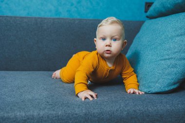 Sarı takım elbiseli sevimli, sarı saçlı küçük bir çocuk kanepede oturuyor. Bebekler için kıyafetler..