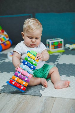 10 aylık sarı saçlı bir çocuk evde yerde oturup oyuncaklarla oynuyor. Bebekler için eğitici oyuncaklar. Çocuğun kişisel gelişimi.