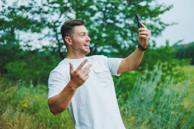 Beyaz tişörtlü bir adam doğada duruyor ve telefonda konuşuyor. Açık hava eğlencesi.